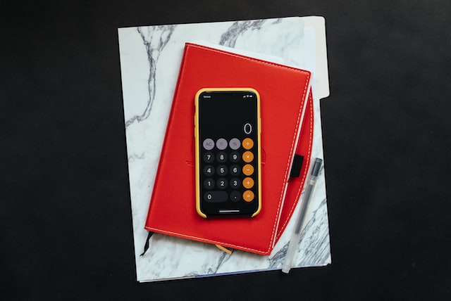 A stack that include a file folder, red notebook, and iphone open to the calculator app